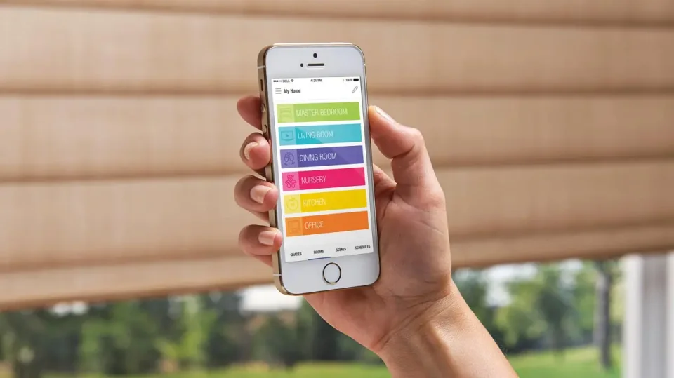 Hand holding a phone beside motorized shades, illustrating a shade control app